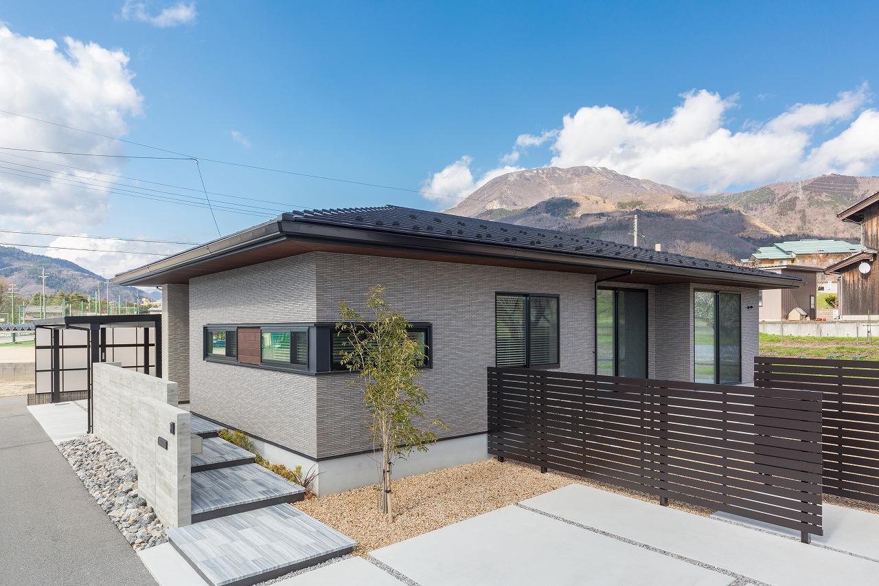 低い切妻屋根の平屋の外観と山並み・木製フェンスのアプローチ